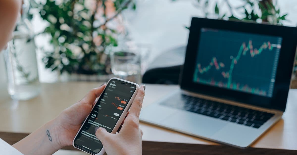 Geschäftsfrau prüft Finanzdaten auf dem Smartphone neben Laptop mit Diagrammen am Schreibtisch.