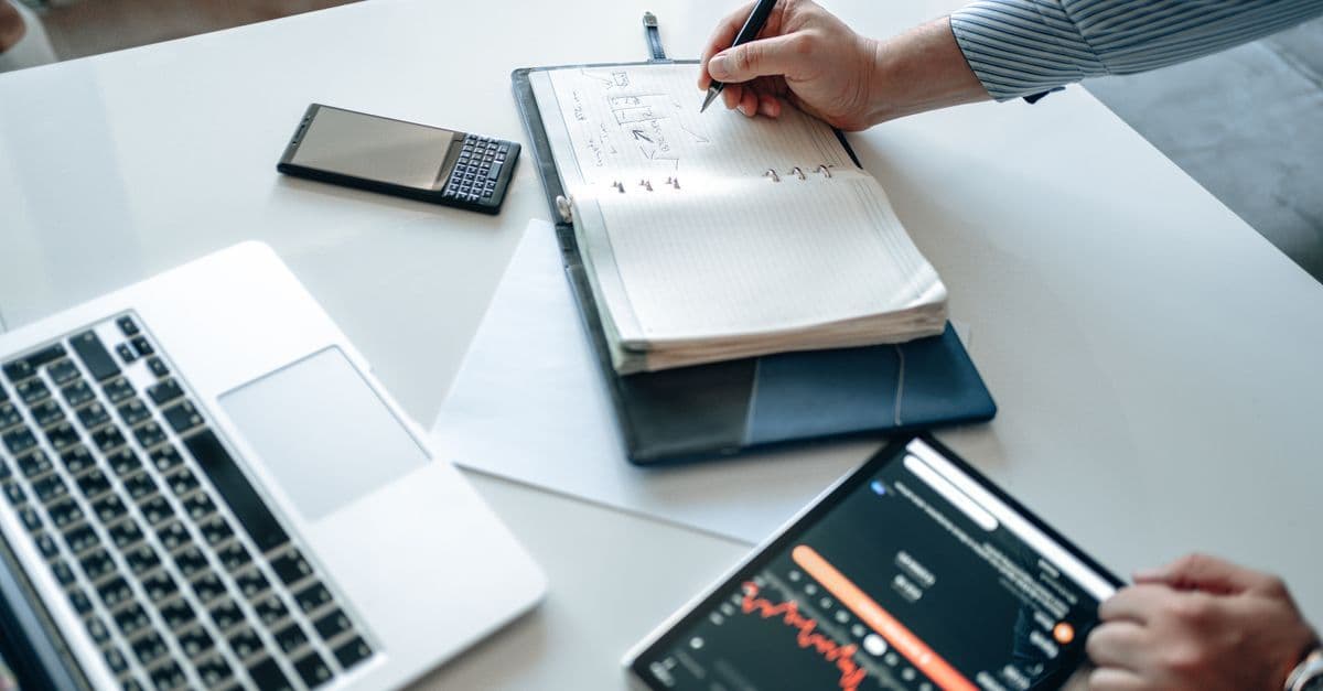 Person analysiert Finanzdaten auf Laptop und Tablet, daneben liegt ein geöffnetes Notizbuch.