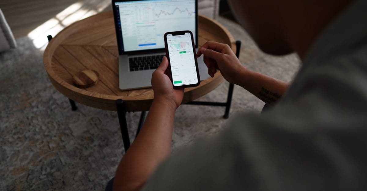 Mann handelt online mit Aktien per Smartphone und Laptop in Innenraumumgebung.