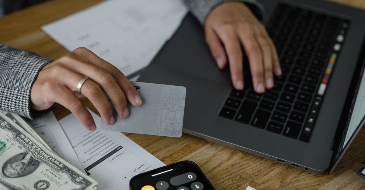 Person zahlt mit Kreditkarte am Laptop, daneben Bargeld und Taschenrechner auf dem Tisch.