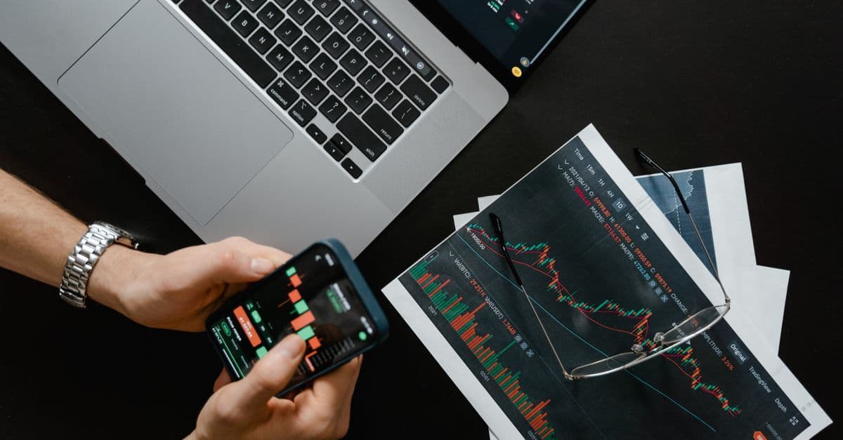 Hände mit Smartphone neben Laptop mit Aktiencharts beim digitalen Handel