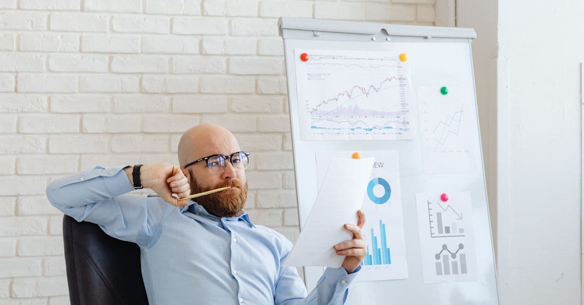Geschäftsmann mit Brille betrachtet nachdenklich Finanzdiagramme vor einem Whiteboard mit Grafiken.