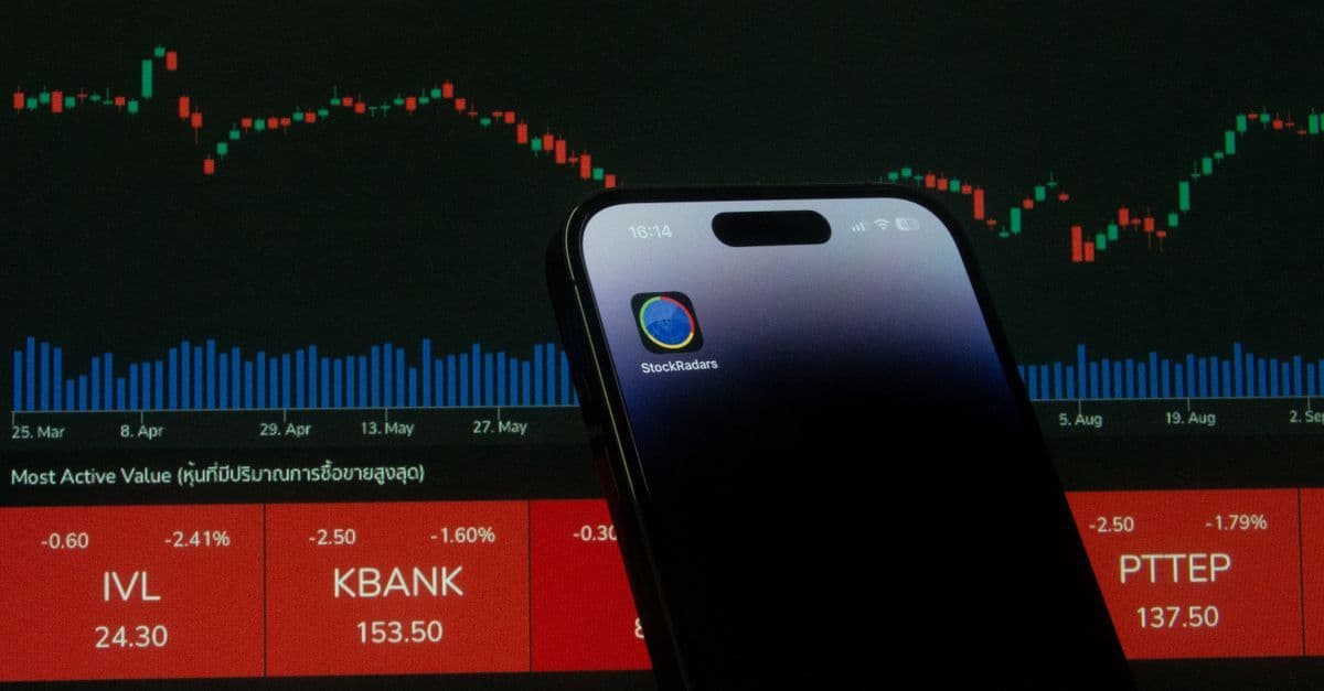 Mobiltelefon mit geöffneter Handels-App vor einem Bildschirm mit Börsenchart.