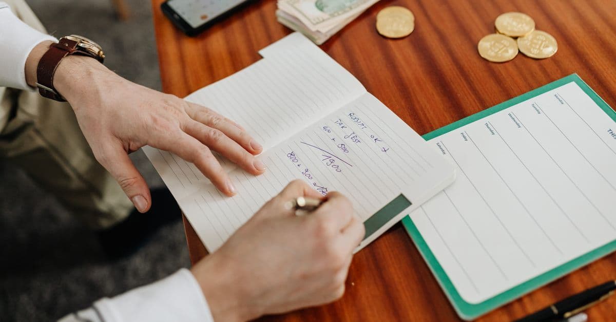 Hände notieren Finanzberechnungen in ein Heft, daneben liegen Geldscheine und Münzen auf dem Tisch.